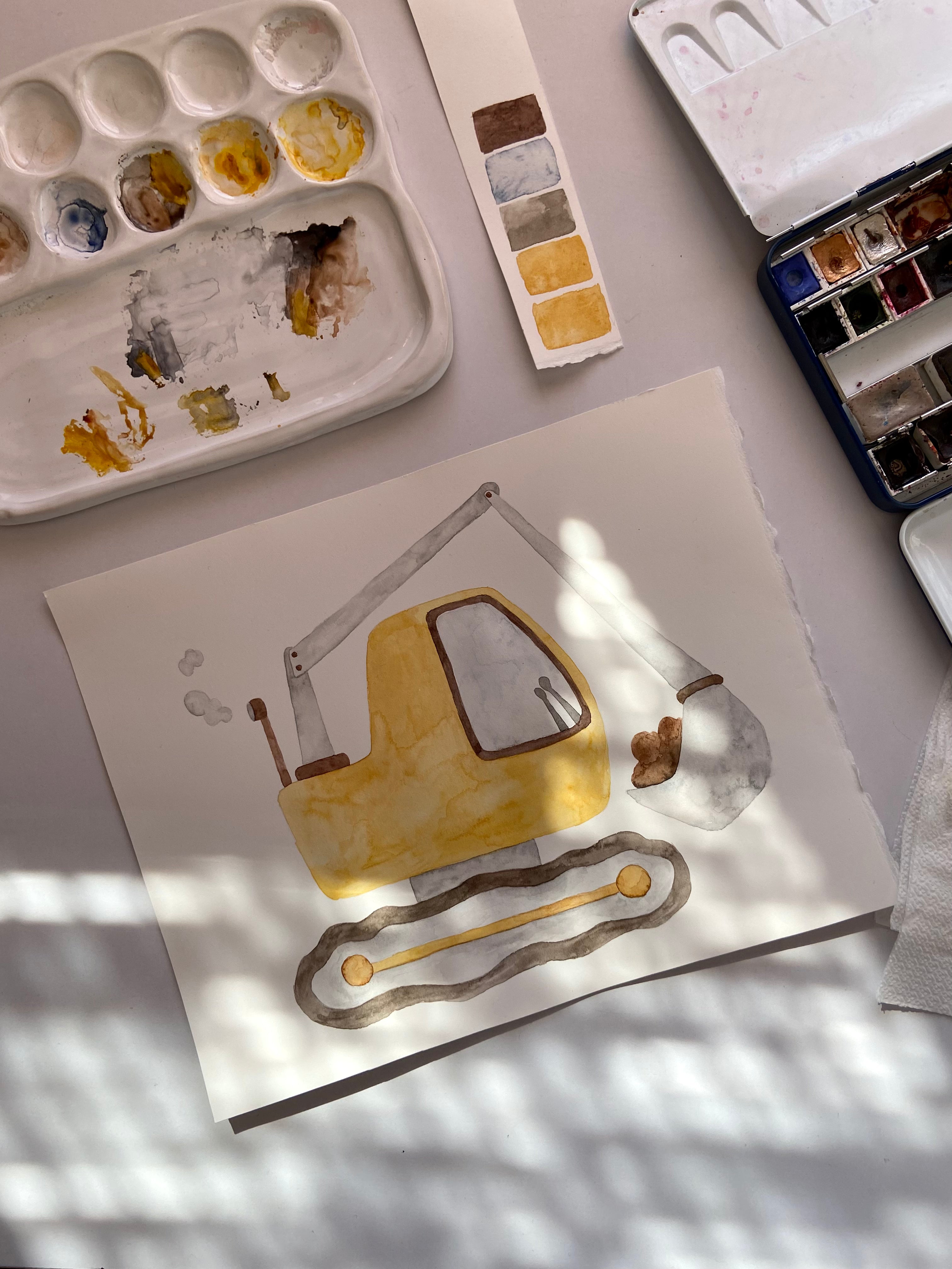 A watercolor painting of a yellow excavator on a white sheet of paper, surrounded by watercolor palettes and paint supplies on a light gray surface with soft window shadows.