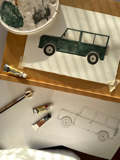 An overhead view of an artist's desk showing a painted dark green vintage car on white paper, alongside a pencil sketch of the same car, paint tubes, a paintbrush, and a paint palette on a wooden surface.